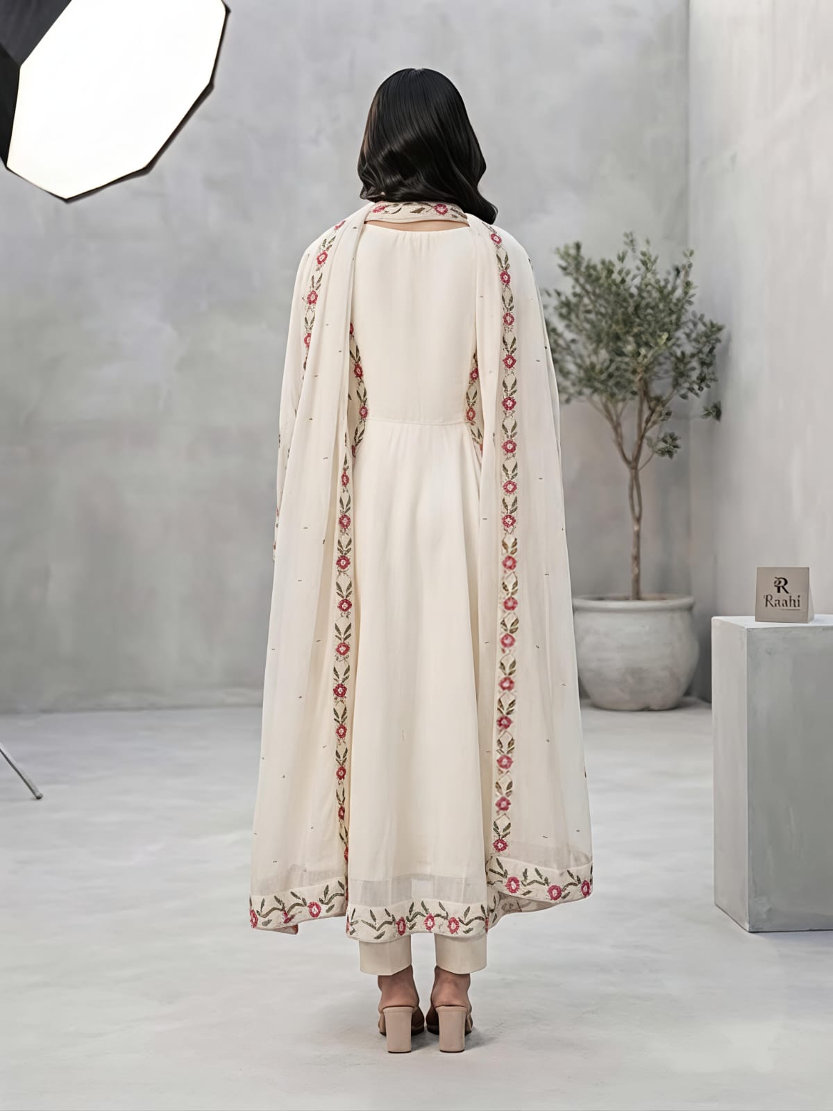A woman models a traditional Indian outfit consisting of a cream-colored Anarkali kurta with red and green floral embroidery, paired with matching trousers and a sheer dupatta, in a studio setting.