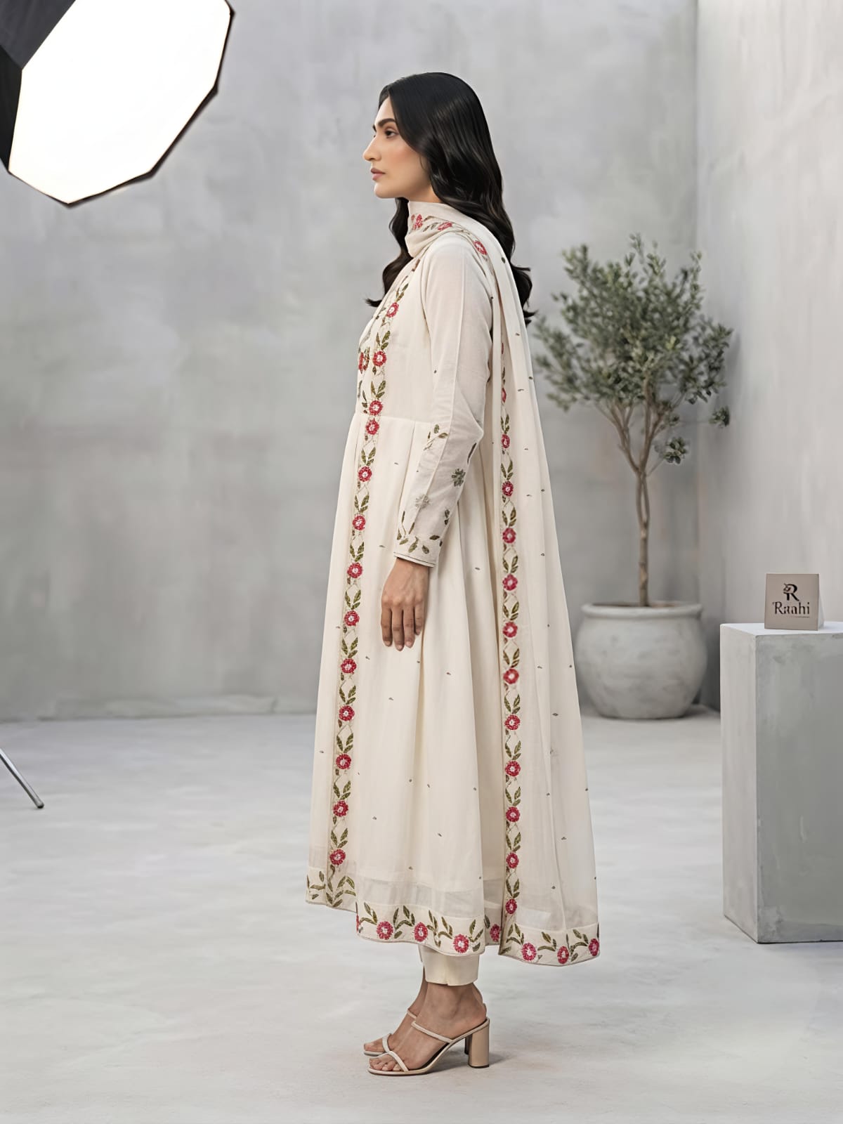 A woman models a traditional Indian outfit consisting of a cream-colored Anarkali kurta with red and green floral embroidery, paired with matching trousers and a sheer dupatta, in a studio setting.