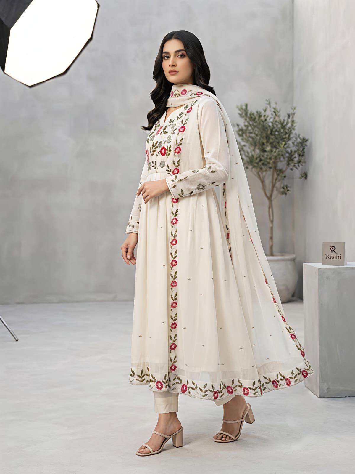A woman models a traditional Indian outfit consisting of a cream-colored Anarkali kurta with red and green floral embroidery, paired with matching trousers and a sheer dupatta, in a studio setting.