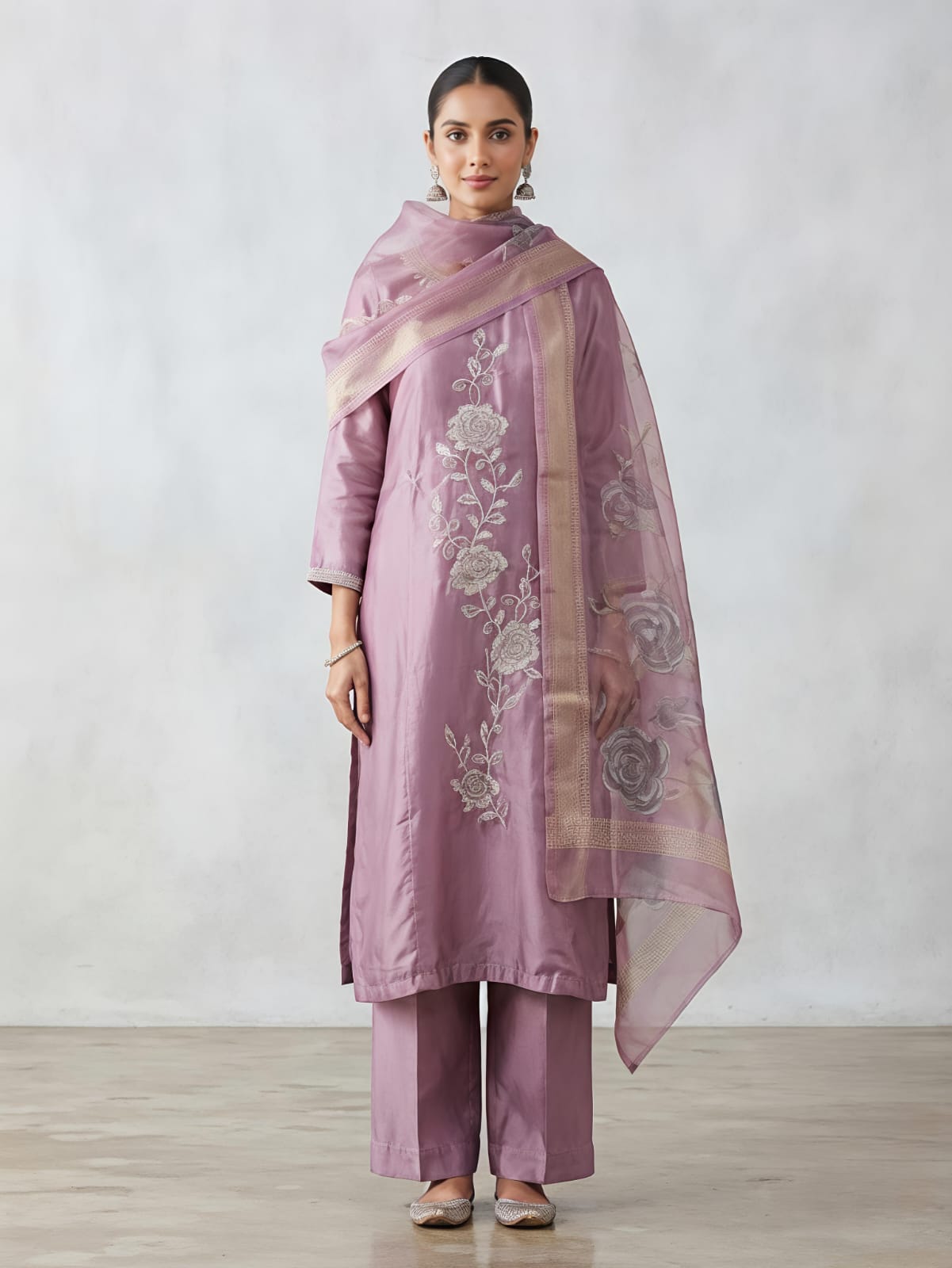 A woman models a lavender purple three-piece traditional outfit consisting of a kurta with floral embroidery, matching trousers, and a sheer dupatta draped over her shoulder, set against a neutral studio background.