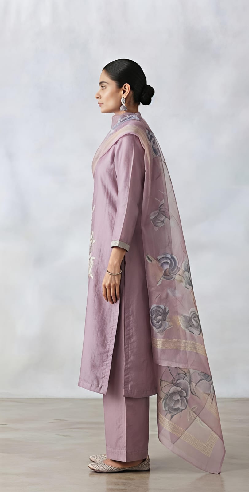 A woman models a lavender purple three-piece traditional outfit consisting of a kurta with floral embroidery, matching trousers, and a sheer dupatta draped over her shoulder, set against a neutral studio background.