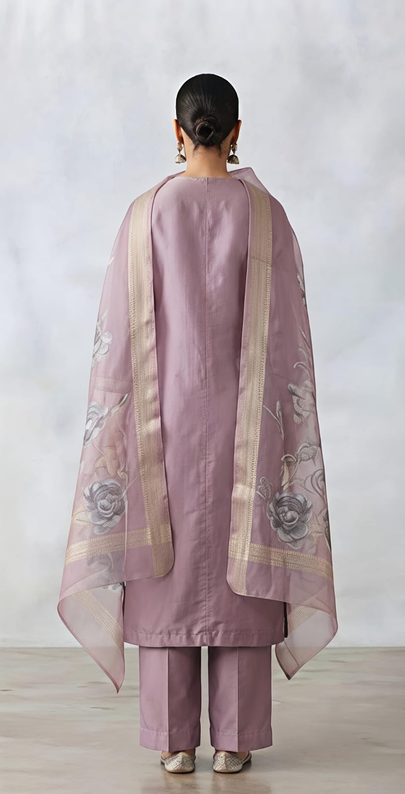 A woman models a lavender purple three-piece traditional outfit consisting of a kurta with floral embroidery, matching trousers, and a sheer dupatta draped over her shoulder, set against a neutral studio background.
