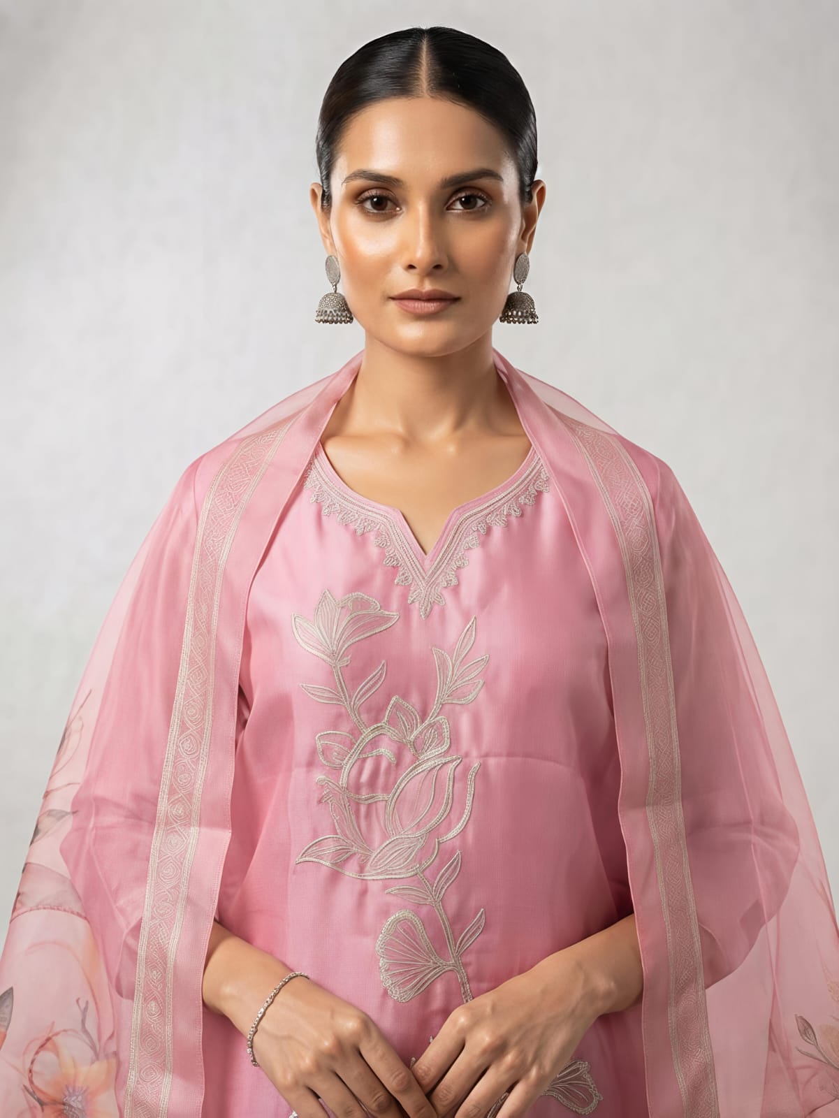 A woman stands in a studio wearing a pink traditional outfit with matching pants and a sheer floral dupatta. The set has light pink floral embroidery on the kurta and pink, orange, and beige floral motifs on the dupatta.