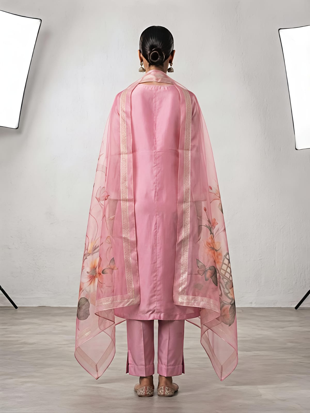 A woman stands in a studio wearing a pink traditional outfit with matching pants and a sheer floral dupatta. The set has light pink floral embroidery on the kurta and pink, orange, and beige floral motifs on the dupatta.