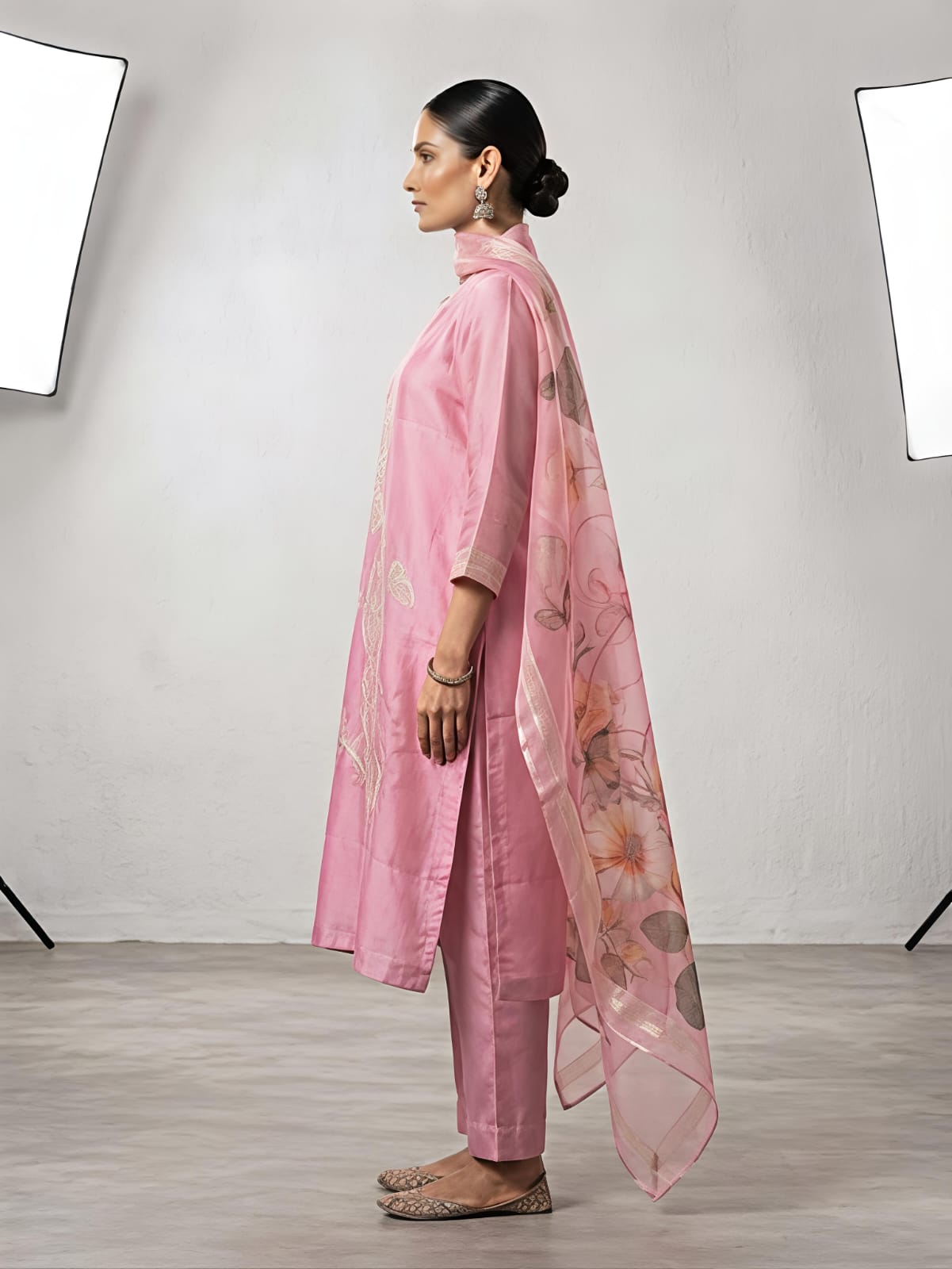 A woman stands in a studio wearing a pink traditional outfit with matching pants and a sheer floral dupatta. The set has light pink floral embroidery on the kurta and pink, orange, and beige floral motifs on the dupatta.
