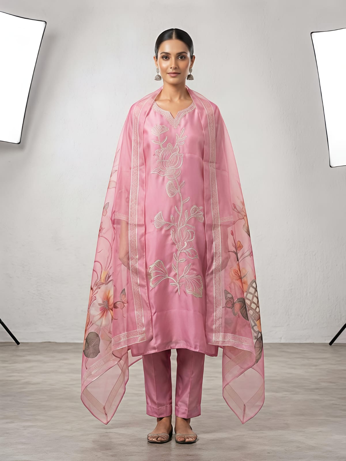 A woman stands in a studio wearing a pink traditional outfit with matching pants and a sheer floral dupatta. The set has light pink floral embroidery on the kurta and pink, orange, and beige floral motifs on the dupatta.