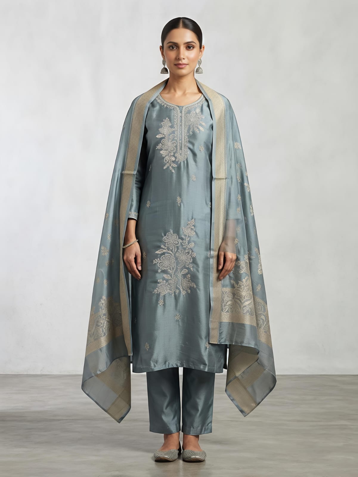 A woman models a traditional three-piece outfit consisting of a dusty blue-green kurta with silver floral embroidery, matching trousers, and a sheer dupatta with beige borders and silver embroidery, set against a neutral studio backdrop.