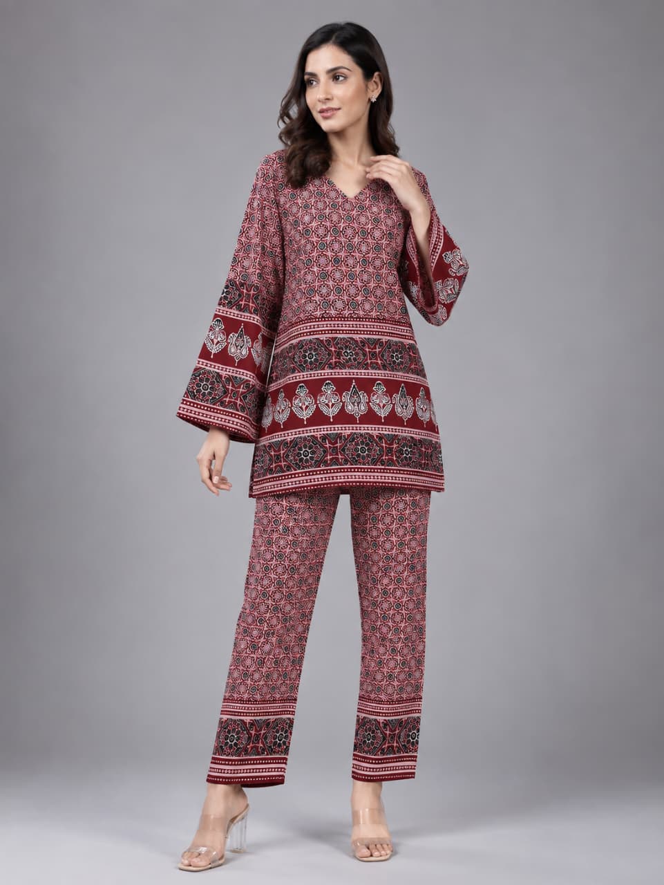 A woman modeling a matching two-piece kurta and trouser set with a red, pink, and black floral block print pattern, set against a solid gray studio background.