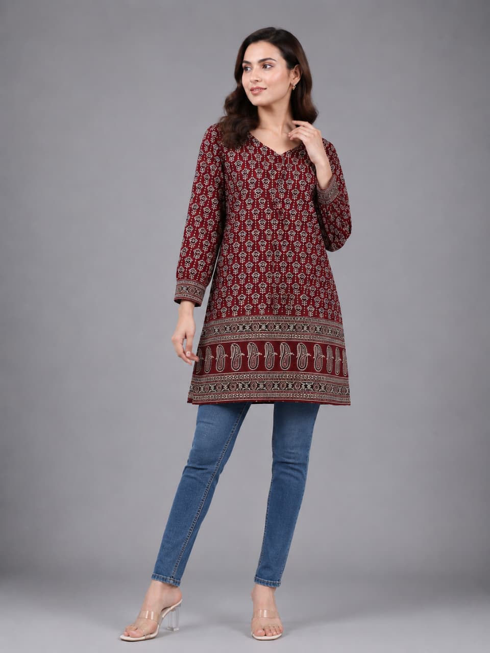 A woman modeling a long-sleeved maroon kurta with a detailed beige and white geometric pattern, paired with blue jeans and clear high-heeled sandals, against a solid gray studio background.