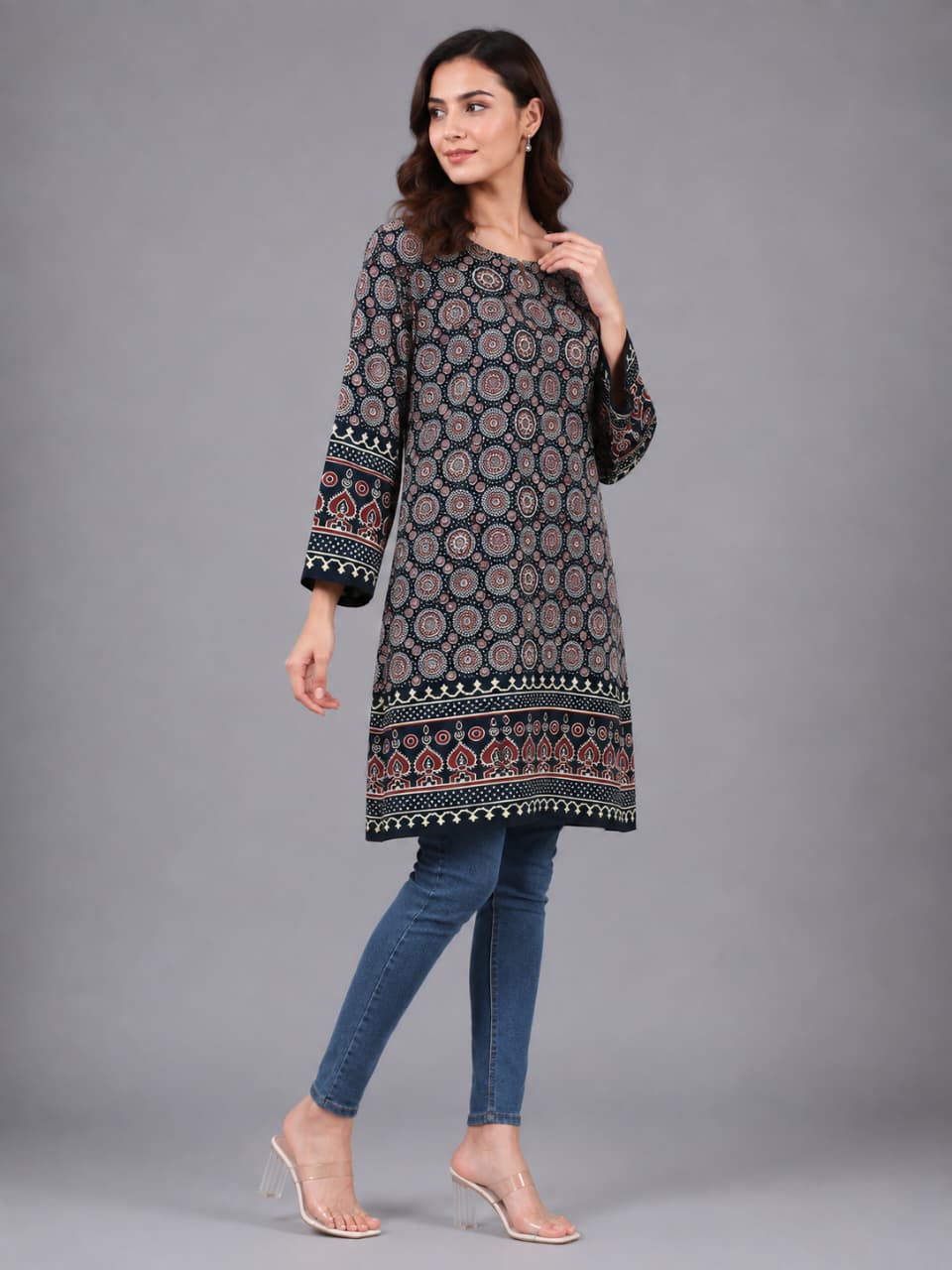 A woman modeling a long-sleeved printed tunic in a dark navy blue base with intricate red, beige, and white geometric patterns, paired with blue skinny jeans and clear high-heeled sandals, against a solid gray studio background.