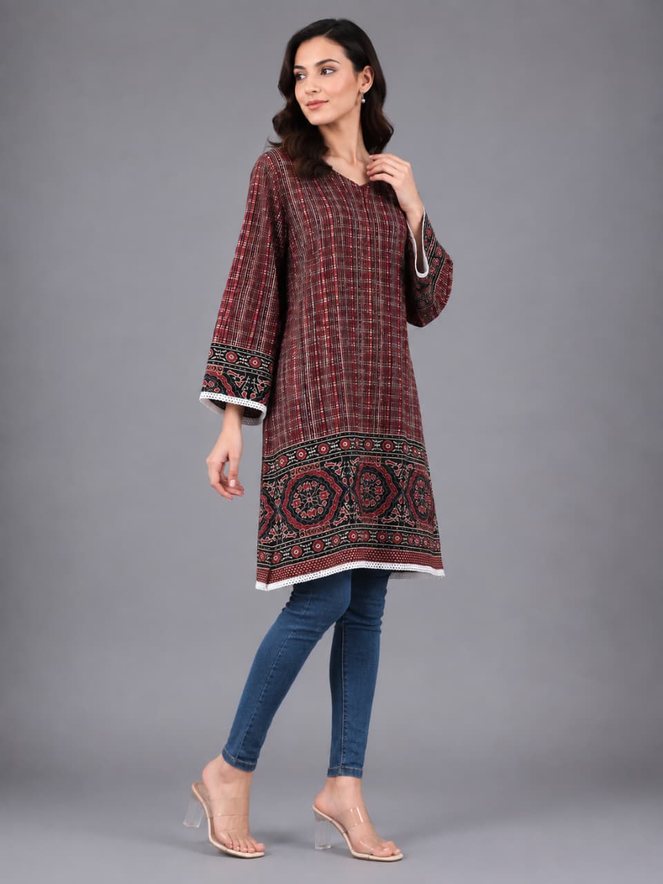 A woman modeling a long-sleeved tunic top with a red and black plaid pattern and intricate geometric embroidery along the hem and cuffs, paired with blue jeans and clear high-heeled sandals, against a solid gray studio background.