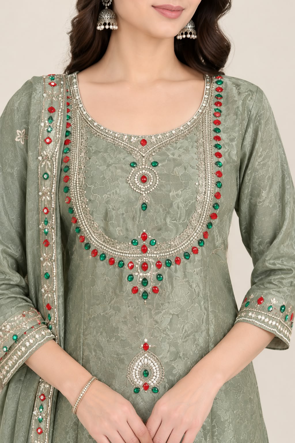A woman models a sage green ethnic salwar kameez suit set with matching dupatta. The kurta features intricate silver zari embroidery and colorful red and green bead embellishments around the neckline and hem, paired with matching sage green trousers and a coordinating dupatta.