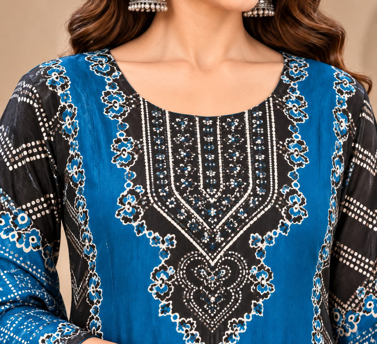 A woman modeling a traditional three-piece salwar kameez suit in a warm beige interior. The outfit features a black kurta with intricate blue and white geometric patterns, paired with black trousers and a matching blue dupatta.
