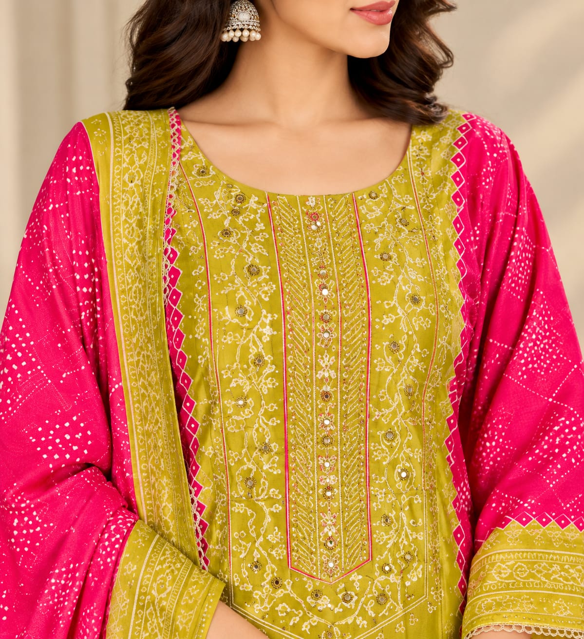 A woman models a traditional Indian salwar kameez suit set in a warm beige indoor setting. The outfit features a lime green kameez with heavy gold embroidery on the chest, matching green trousers, and a vibrant pink dupatta with gold borders and stripes.