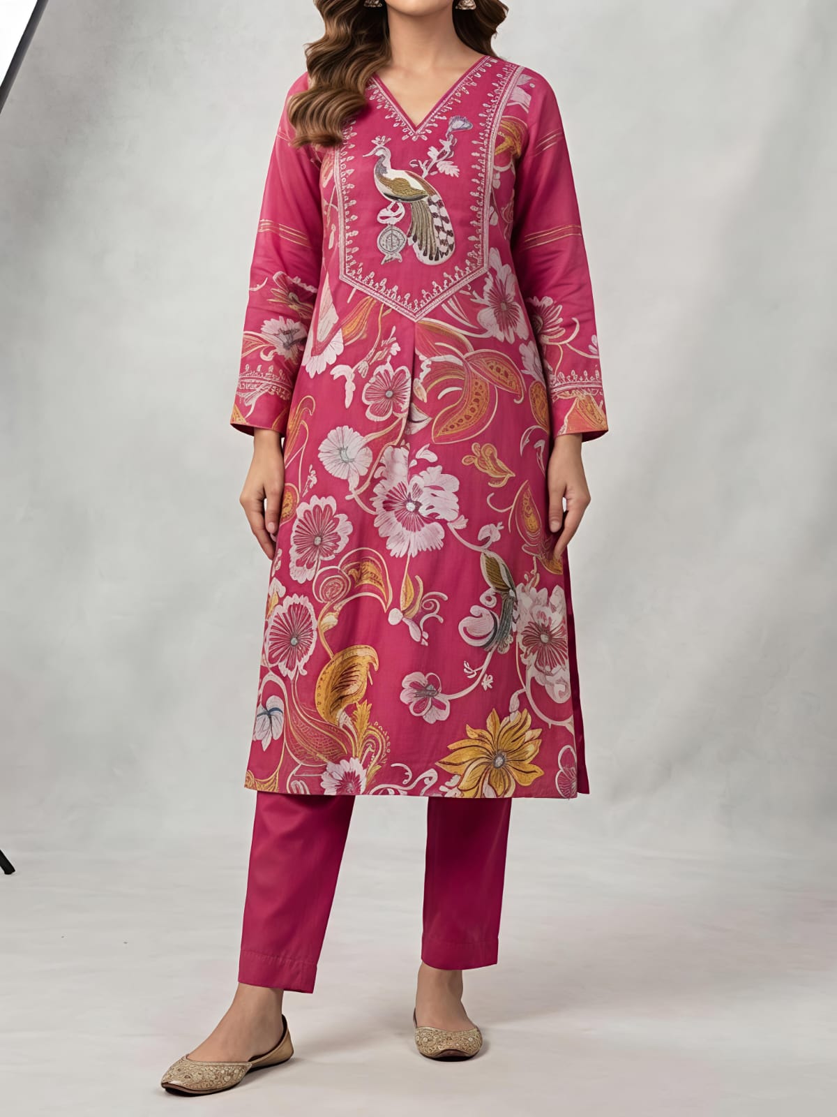 A woman models a bright pink floral printed salwar kameez set with matching trousers and a sheer dupatta, photographed in a studio setting with a softbox light visible on the left.
