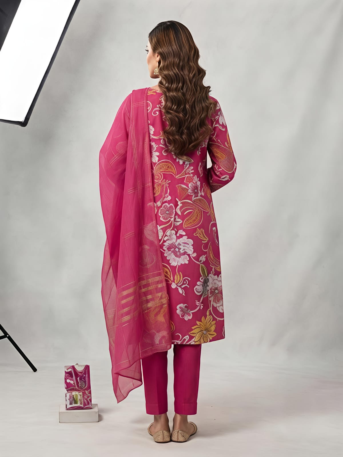 A woman models a bright pink floral printed salwar kameez set with matching trousers and a sheer dupatta, photographed in a studio setting with a softbox light visible on the left.