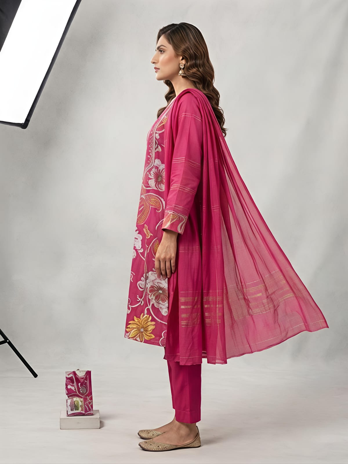 A woman models a bright pink floral printed salwar kameez set with matching trousers and a sheer dupatta, photographed in a studio setting with a softbox light visible on the left.