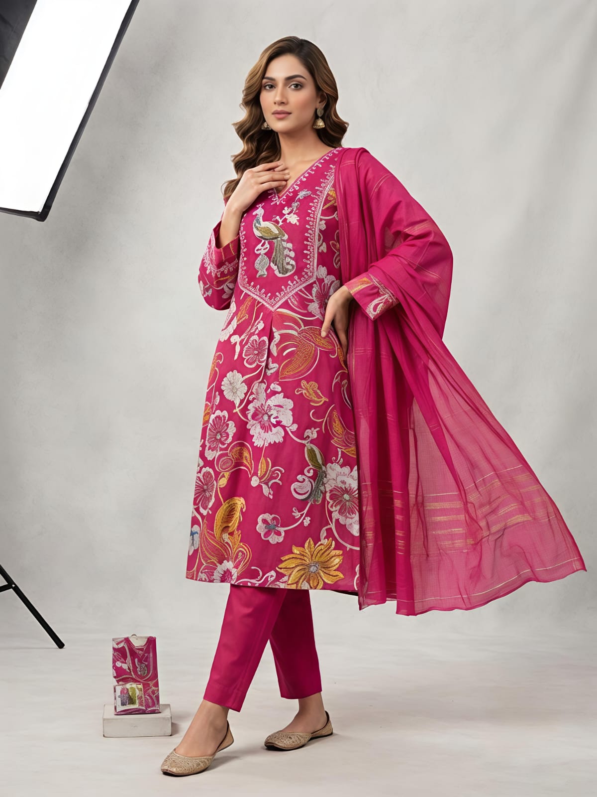 A woman models a bright pink floral printed salwar kameez set with matching trousers and a sheer dupatta, photographed in a studio setting with a softbox light visible on the left.