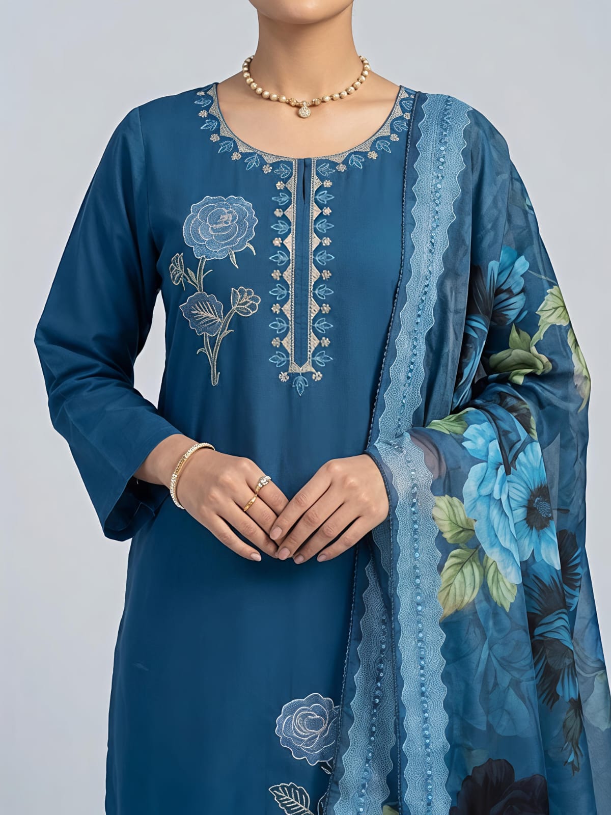 A woman models a traditional blue salwar kameez suit with a floral printed dupatta. The set features a blue long-sleeve kurta with silver embroidery, matching blue trousers, and a sheer blue dupatta decorated with large blue, black, and green floral motifs.