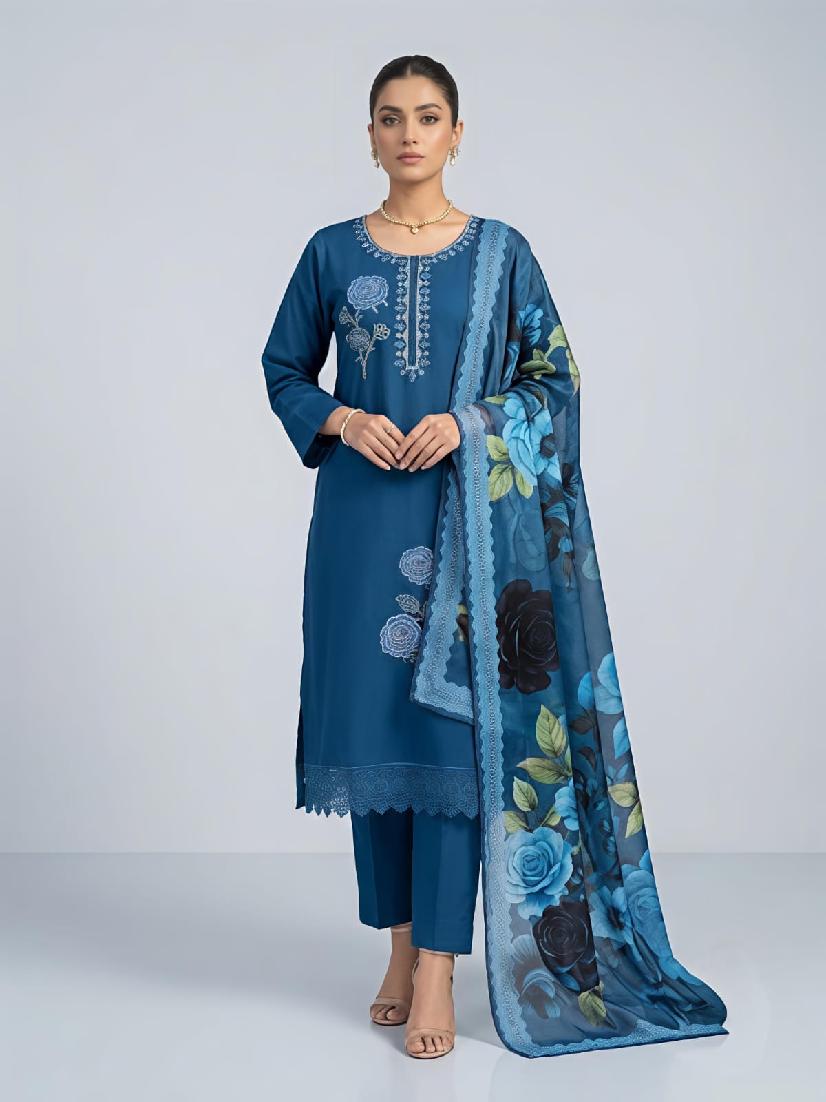 A woman models a traditional blue salwar kameez suit with a floral printed dupatta. The set features a blue long-sleeve kurta with silver embroidery, matching blue trousers, and a sheer blue dupatta decorated with large blue, black, and green floral motifs.