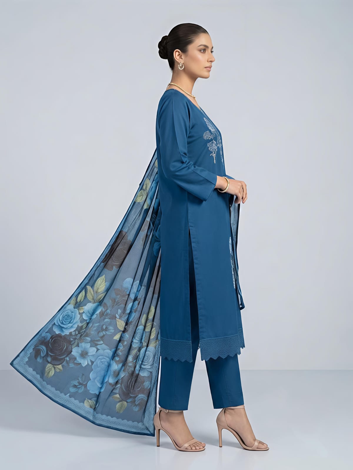 A woman models a traditional blue salwar kameez suit with a floral printed dupatta. The set features a blue long-sleeve kurta with silver embroidery, matching blue trousers, and a sheer blue dupatta decorated with large blue, black, and green floral motifs.