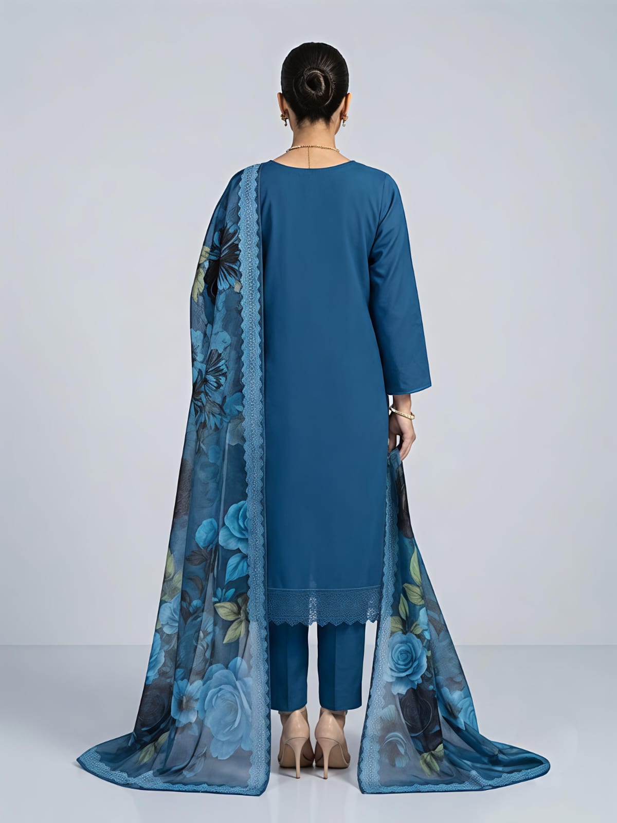 A woman models a traditional blue salwar kameez suit with a floral printed dupatta. The set features a blue long-sleeve kurta with silver embroidery, matching blue trousers, and a sheer blue dupatta decorated with large blue, black, and green floral motifs.