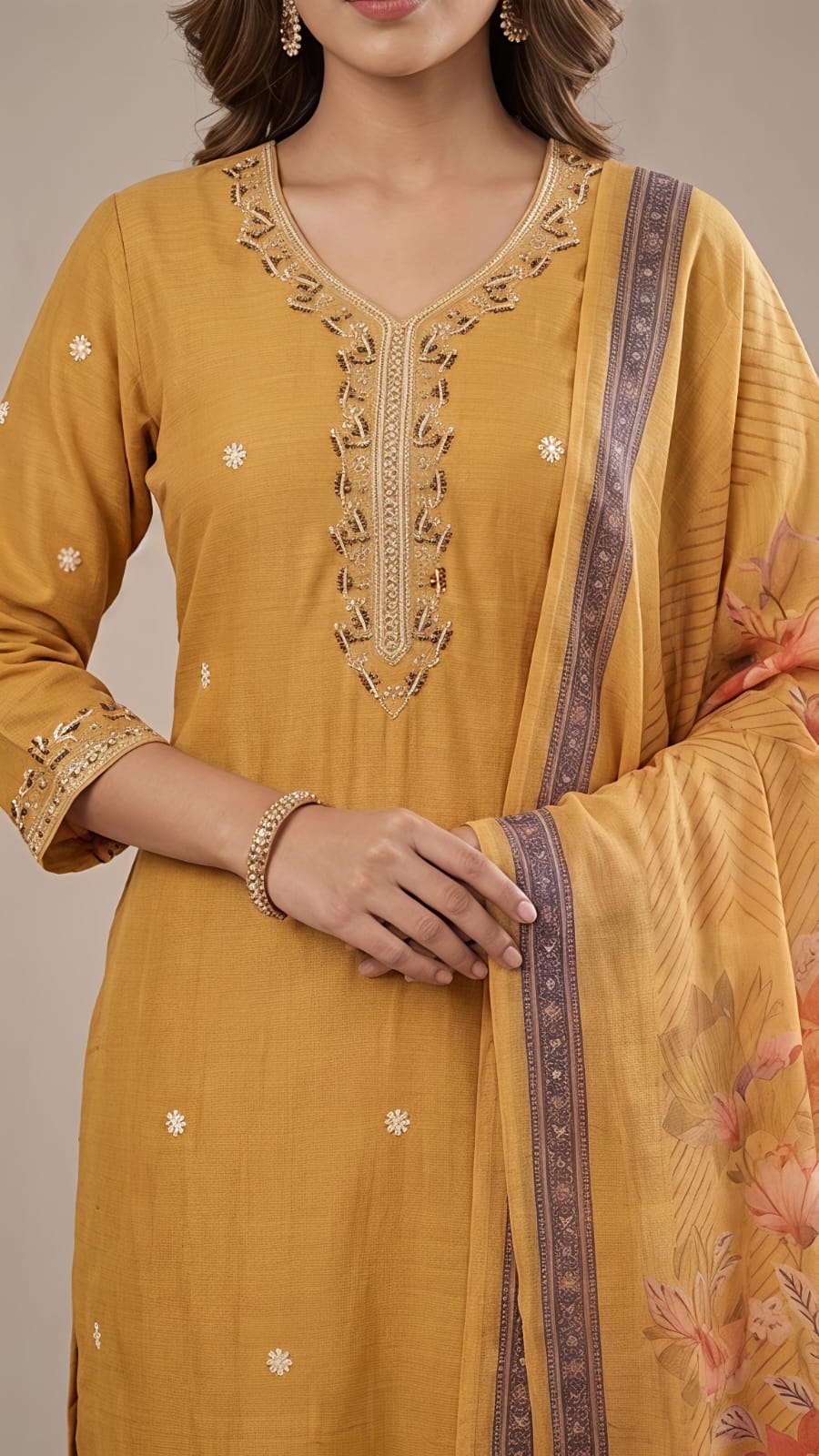 A woman modeling a mustard yellow ethnic suit set with a matching dupatta. The kurta features embroidered details on the neckline and small white dot embellishments, paired with matching trousers and a printed dupatta with a purple border and floral motifs.