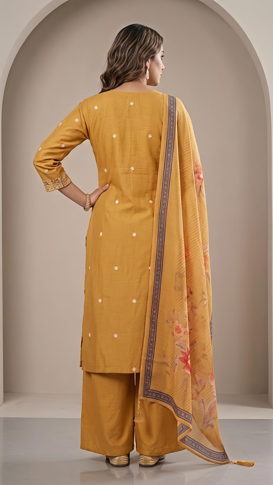 A woman modeling a mustard yellow ethnic suit set with a matching dupatta. The kurta features embroidered details on the neckline and small white dot embellishments, paired with matching trousers and a printed dupatta with a purple border and floral motifs.