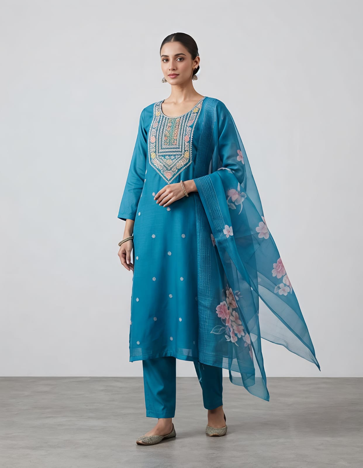 A woman modeling a teal blue Indian ethnic suit set, featuring a kurta with colorful embroidered neckline, matching trousers, and a sheer floral printed dupatta, set against a neutral studio background.