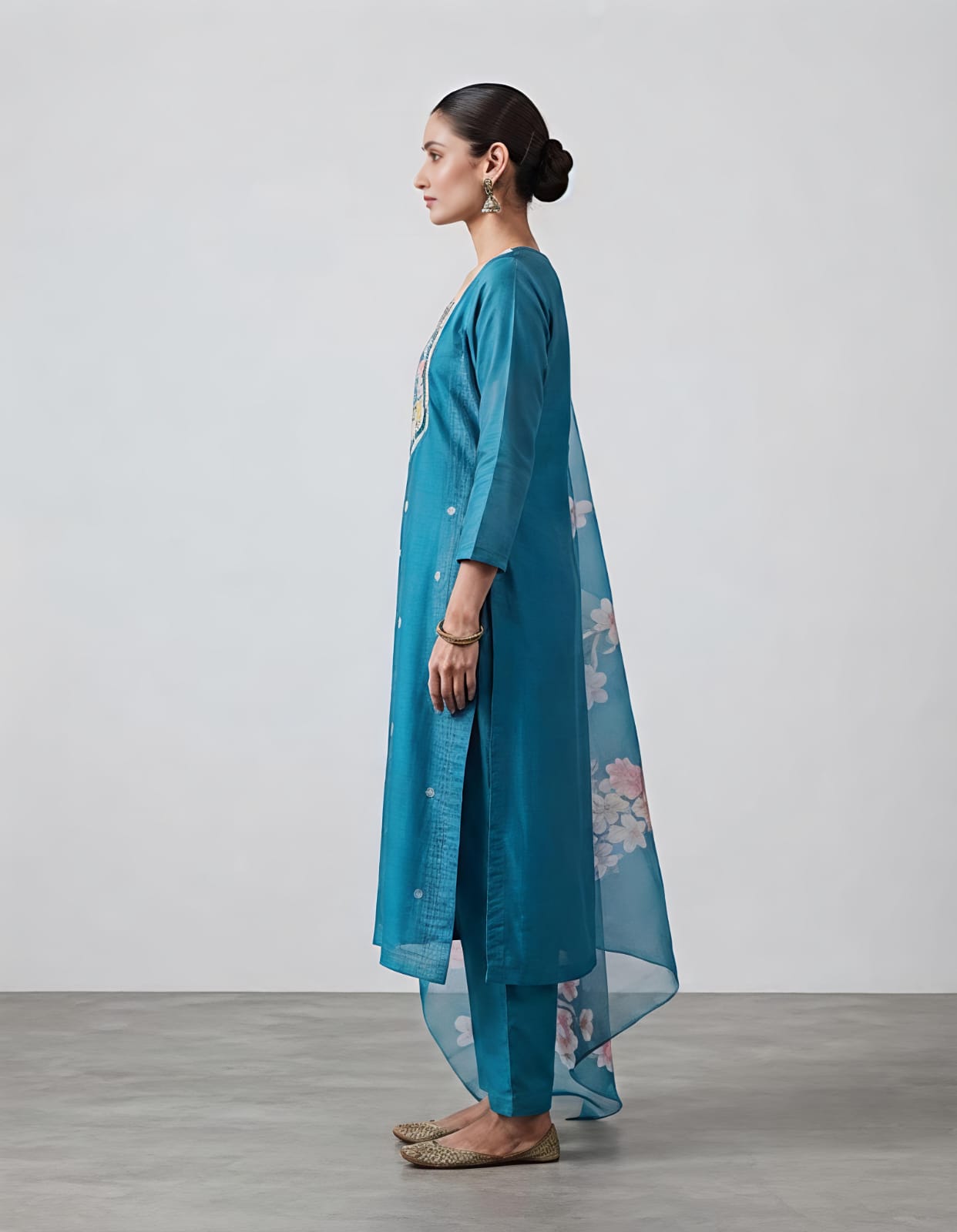 A woman modeling a teal blue Indian ethnic suit set, featuring a kurta with colorful embroidered neckline, matching trousers, and a sheer floral printed dupatta, set against a neutral studio background.