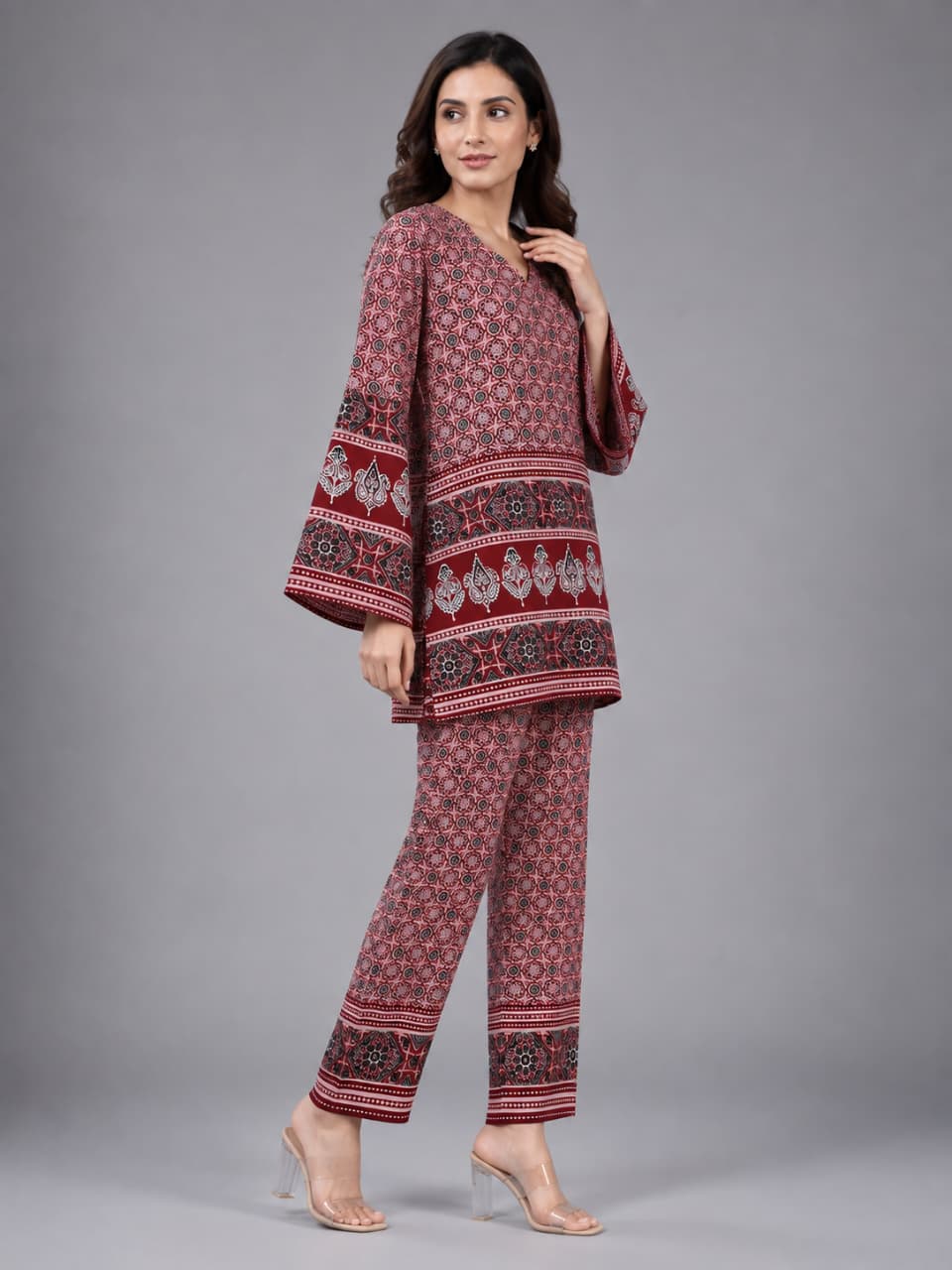 A woman modeling a matching two-piece kurta and trouser set with a red, pink, and black floral block print pattern, set against a solid gray studio background.