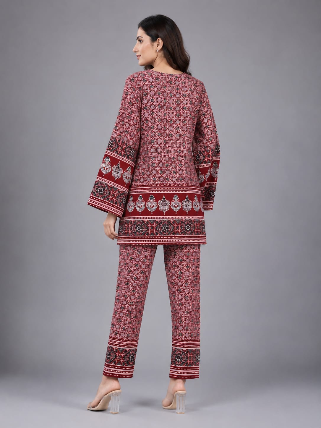 A woman modeling a matching two-piece kurta and trouser set with a red, pink, and black floral block print pattern, set against a solid gray studio background.