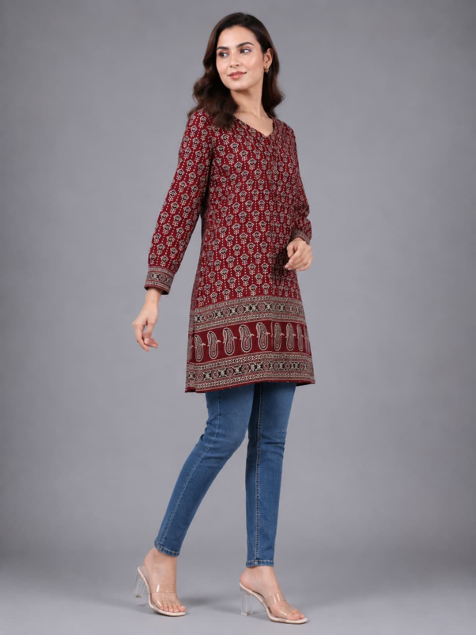 A woman modeling a long-sleeved maroon kurta with a detailed beige and white geometric pattern, paired with blue jeans and clear high-heeled sandals, against a solid gray studio background.