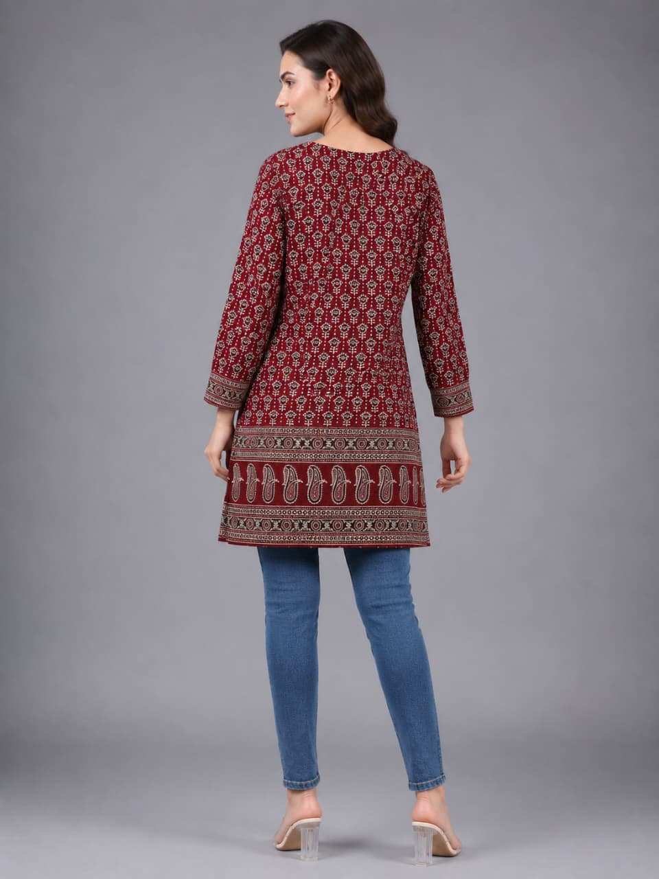 A woman modeling a long-sleeved maroon kurta with a detailed beige and white geometric pattern, paired with blue jeans and clear high-heeled sandals, against a solid gray studio background.