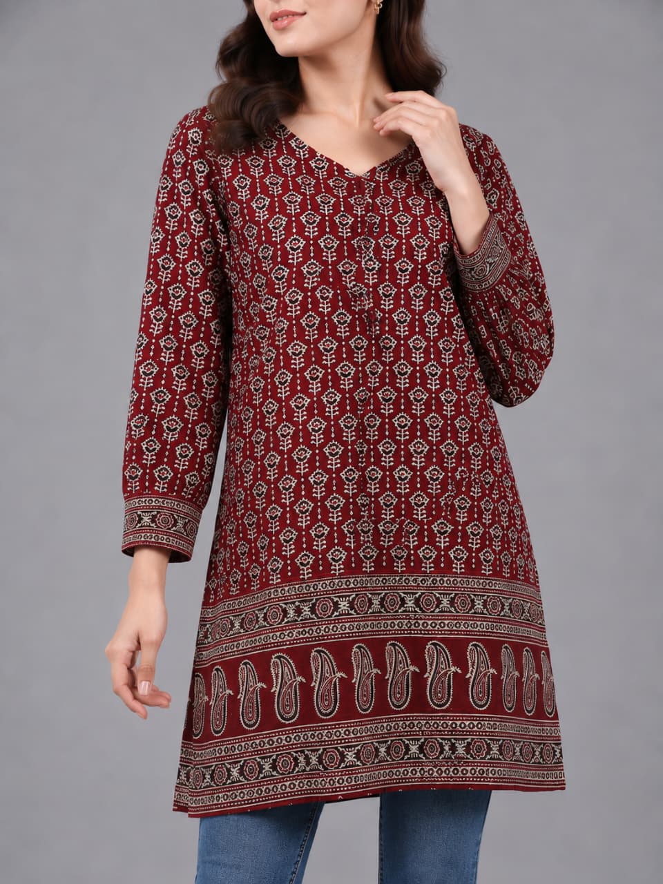 A woman modeling a long-sleeved maroon kurta with a detailed beige and white geometric pattern, paired with blue jeans and clear high-heeled sandals, against a solid gray studio background.