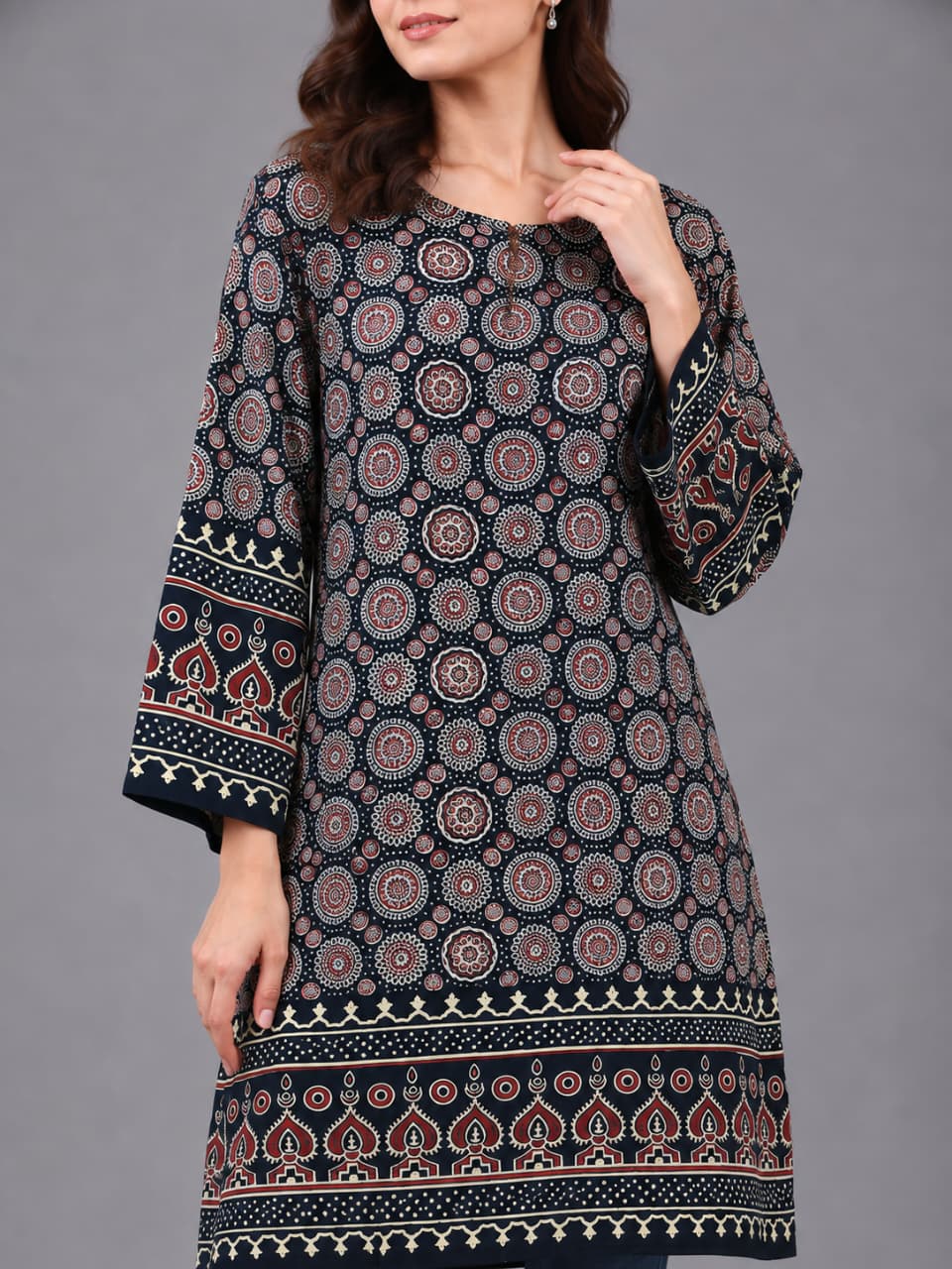 A woman modeling a long-sleeved printed tunic in a dark navy blue base with intricate red, beige, and white geometric patterns, paired with blue skinny jeans and clear high-heeled sandals, against a solid gray studio background