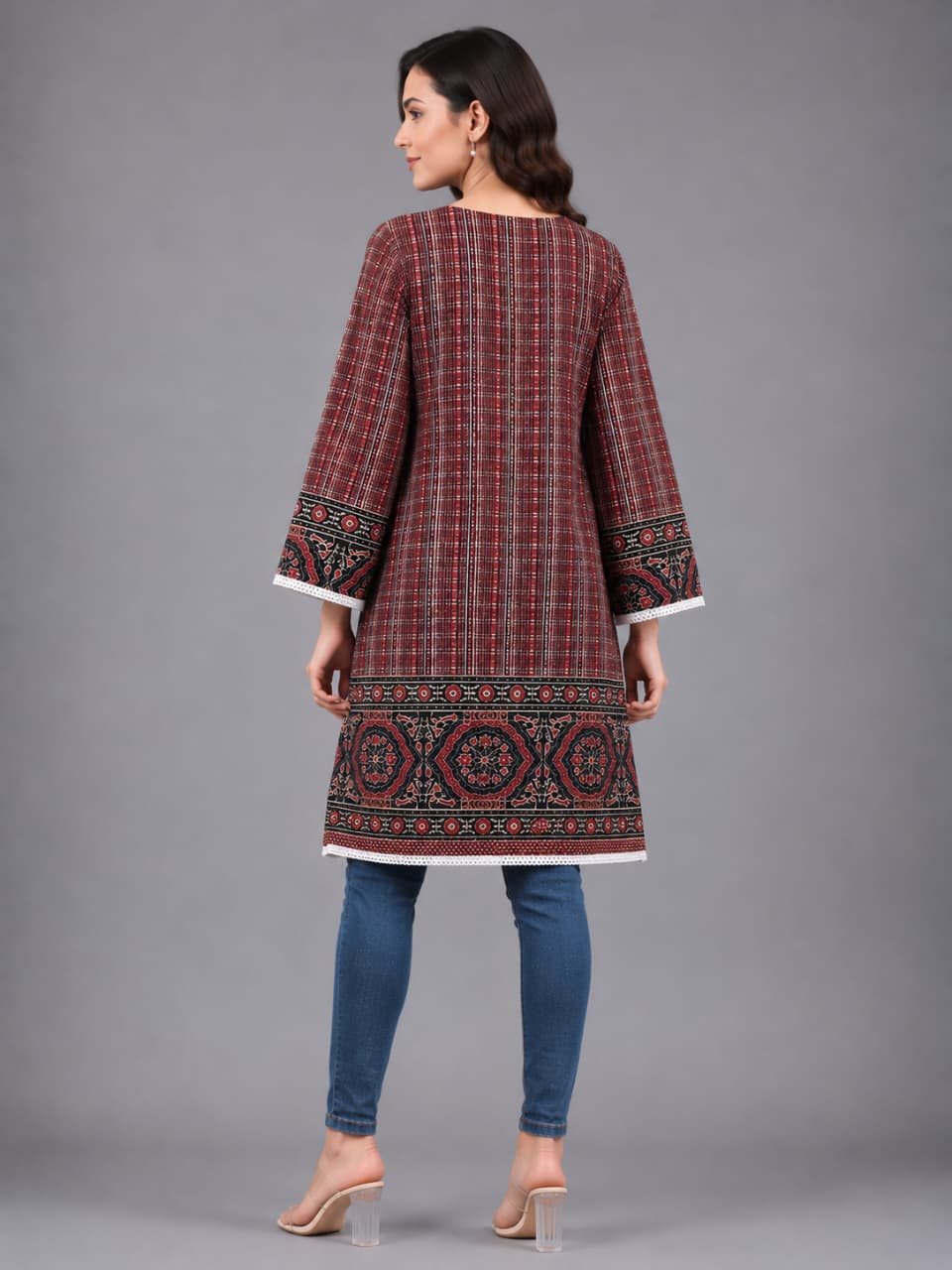A woman modeling a long-sleeved tunic top with a red and black plaid pattern and intricate geometric embroidery along the hem and cuffs, paired with blue jeans and clear high-heeled sandals, against a solid gray studio background