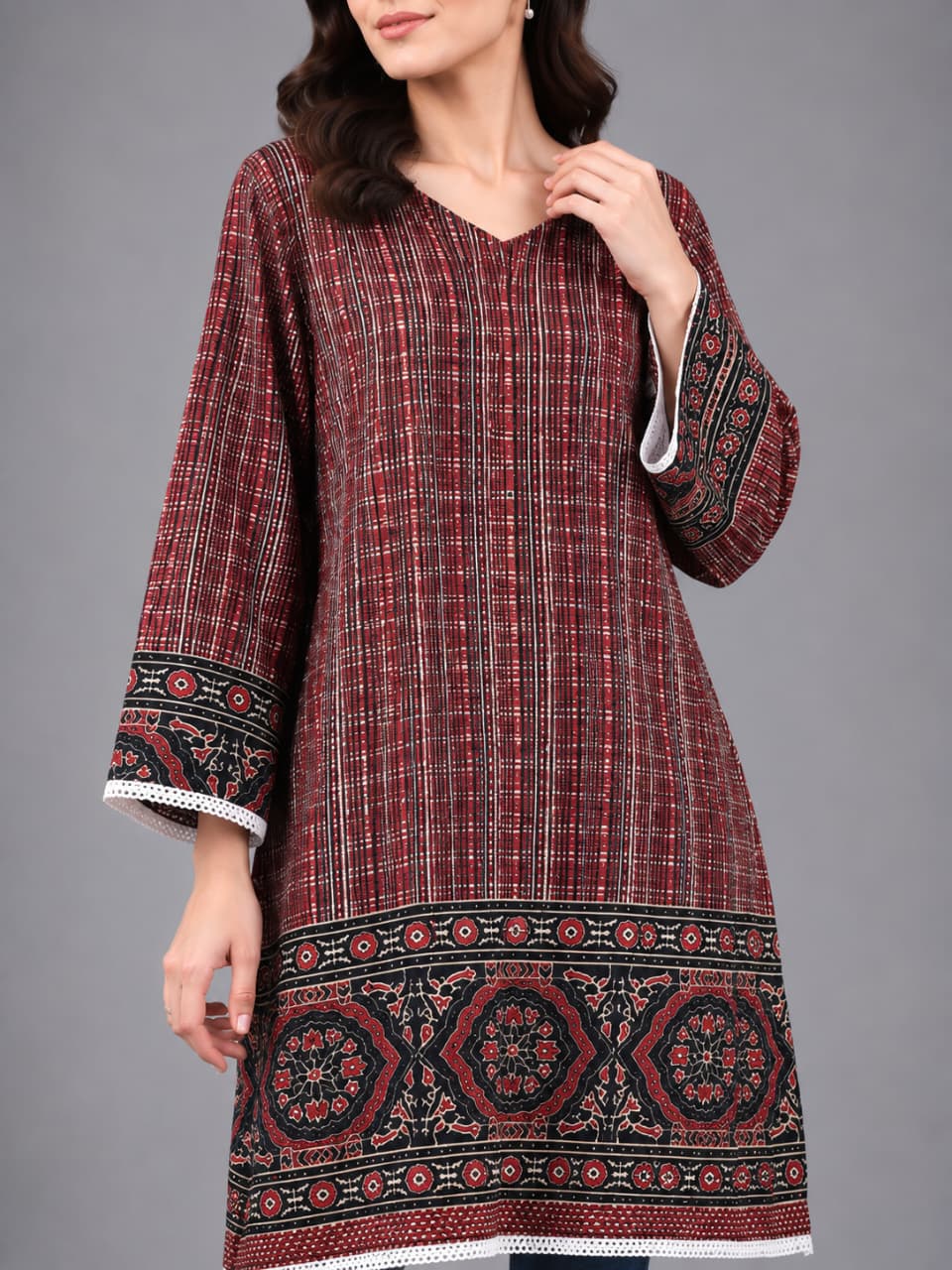 A woman modeling a long-sleeved tunic top with a red and black plaid pattern and intricate geometric embroidery along the hem and cuffs, paired with blue jeans and clear high-heeled sandals, against a solid gray studio background