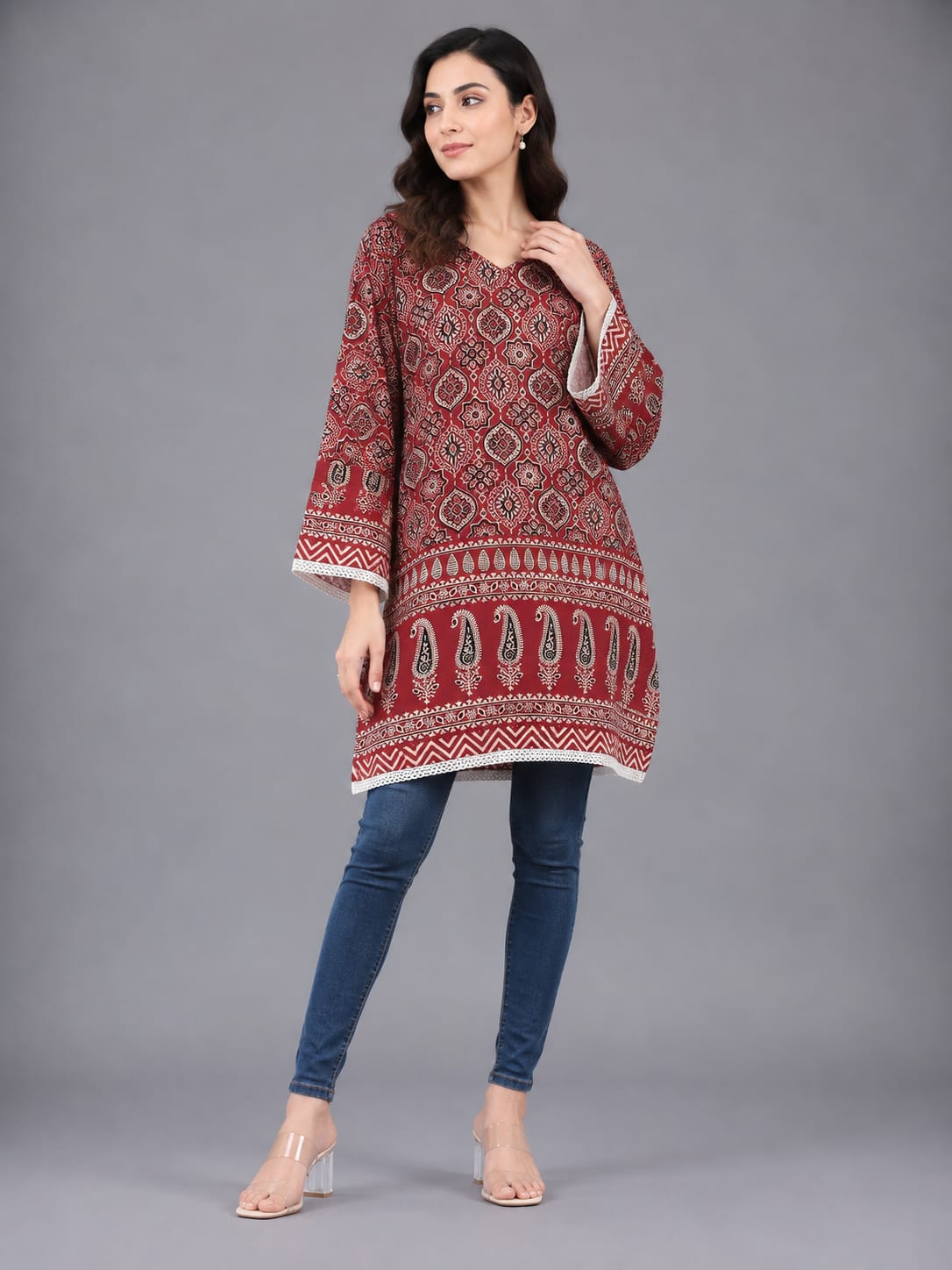 A woman modeling a red and white patterned cotton kurta with blue skinny jeans and clear high-heeled sandals, standing against a solid gray studio background.