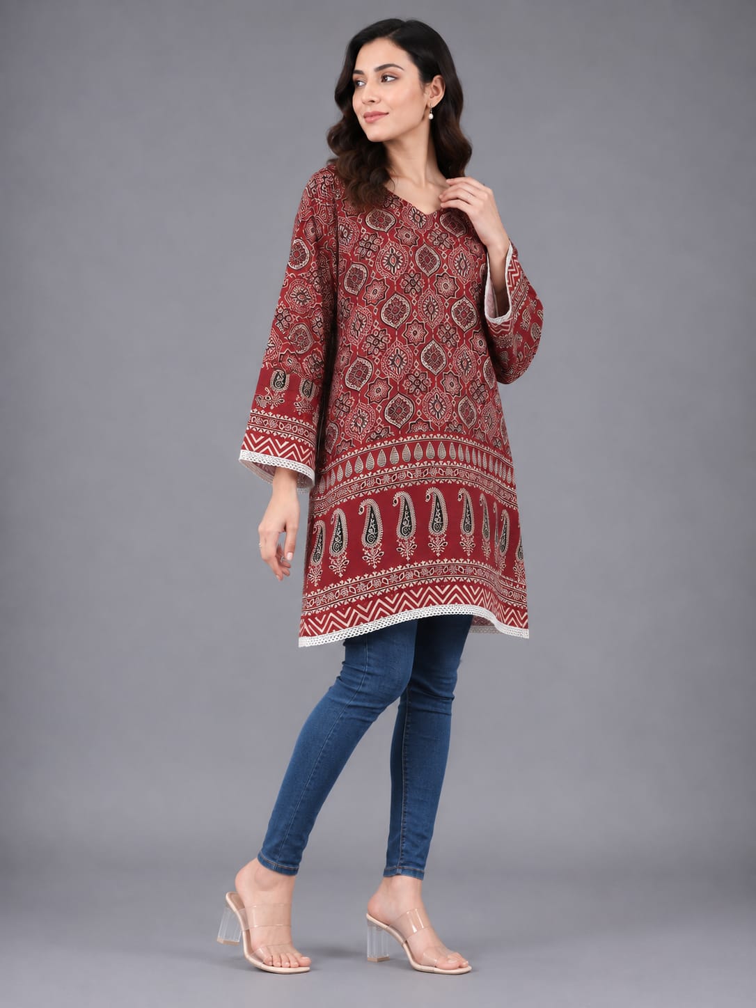 A woman modeling a red and white patterned cotton kurta with blue skinny jeans and clear high-heeled sandals, standing against a solid gray studio background.