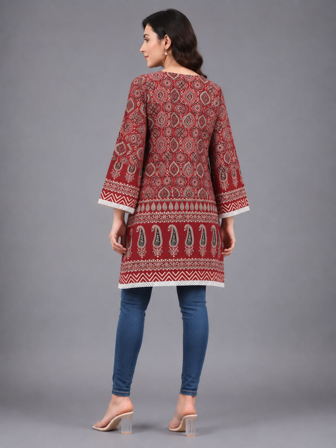 A woman modeling a red and white patterned cotton kurta with blue skinny jeans and clear high-heeled sandals, standing against a solid gray studio background.
