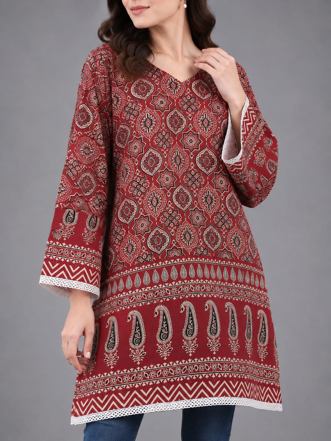 A woman modeling a red and white patterned cotton kurta with blue skinny jeans and clear high-heeled sandals, standing against a solid gray studio background.
