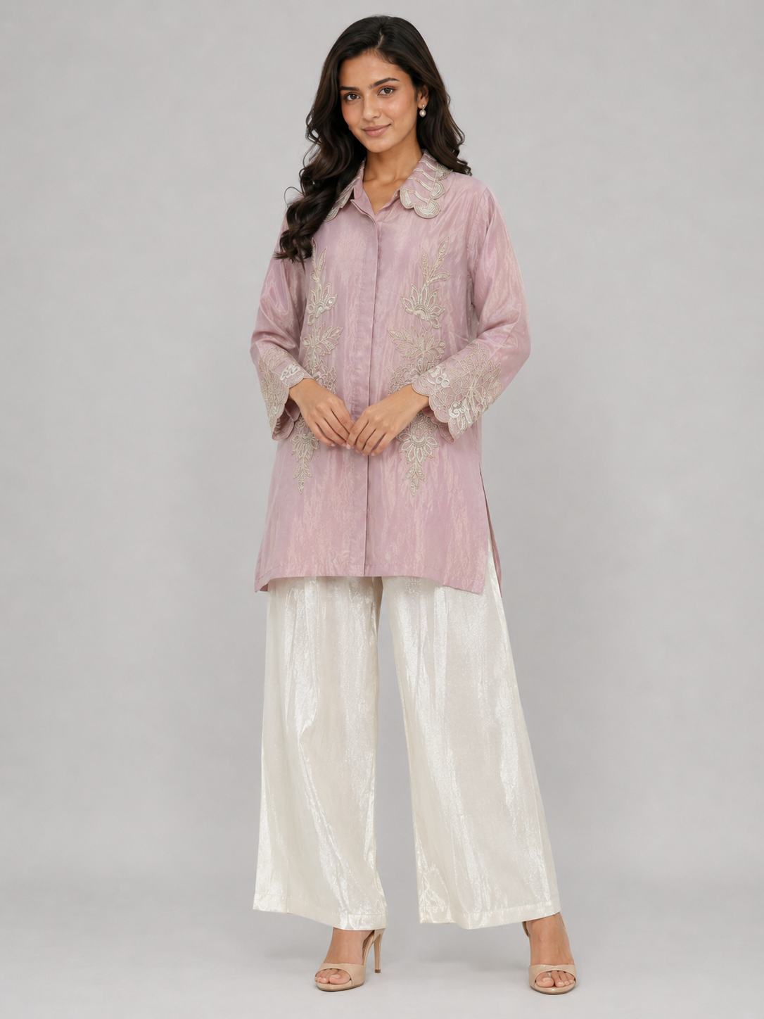 A woman modeling a light pink embroidered kurta paired with white satin palazzo pants and nude heels, set against a solid gray studio background.