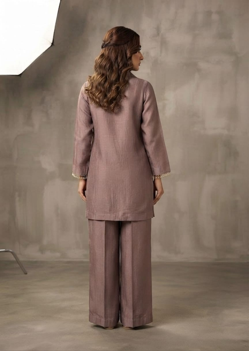 A woman modeling a dusty mauve two-piece kurta and trouser set with floral embroidery on the chest, standing against a textured gray studio backdrop.
