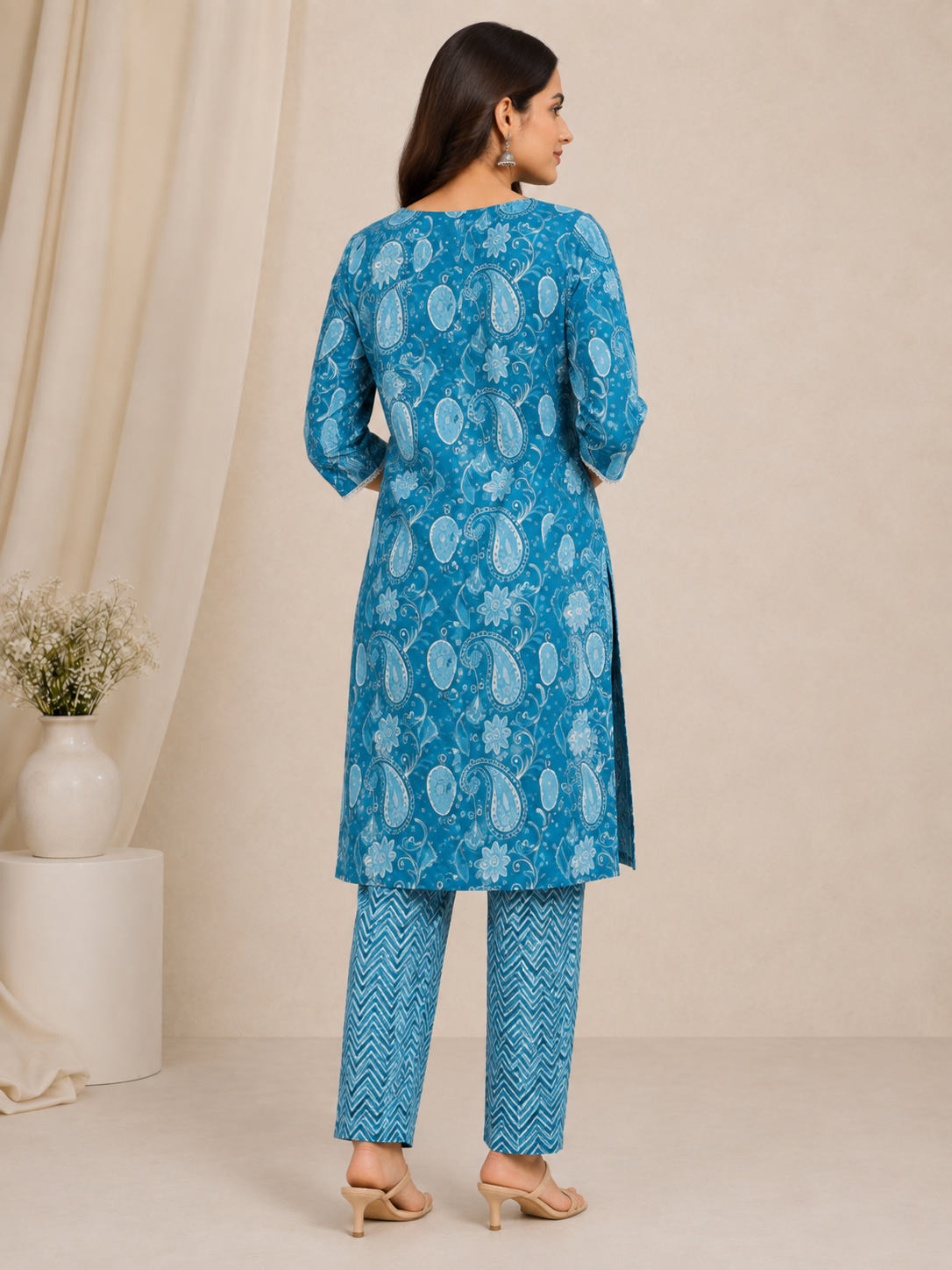 A woman modeling a blue printed kurta and matching pants set with beige heeled sandals, standing in a studio with a neutral beige backdrop and a small vase of flowers on the left.