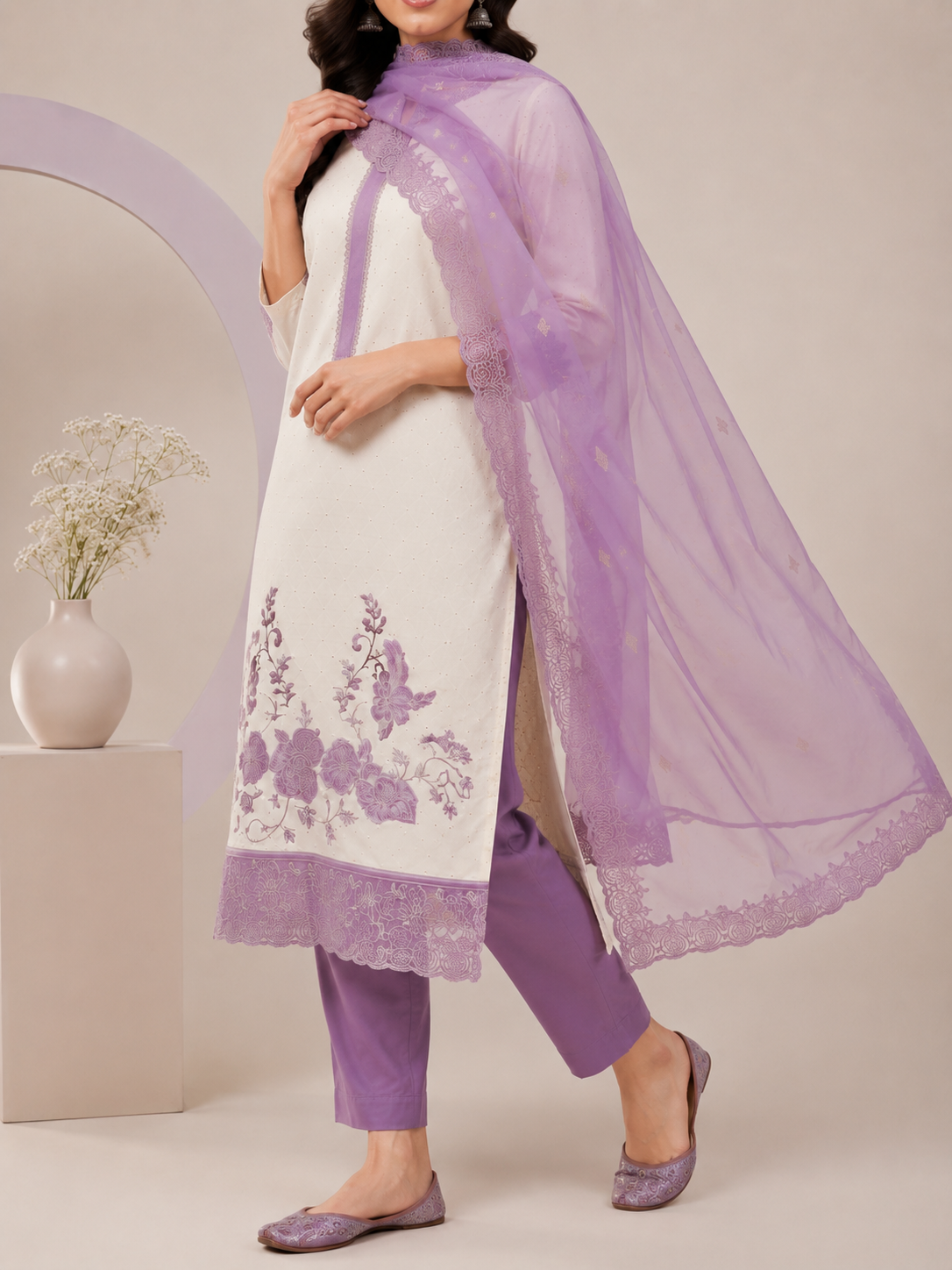 A woman models a traditional Indian salwar kameez suit set. The outfit features an off-white kurta with purple floral embroidery near the hem, paired with matching purple trousers and a sheer lavender dupatta draped over her shoulder.