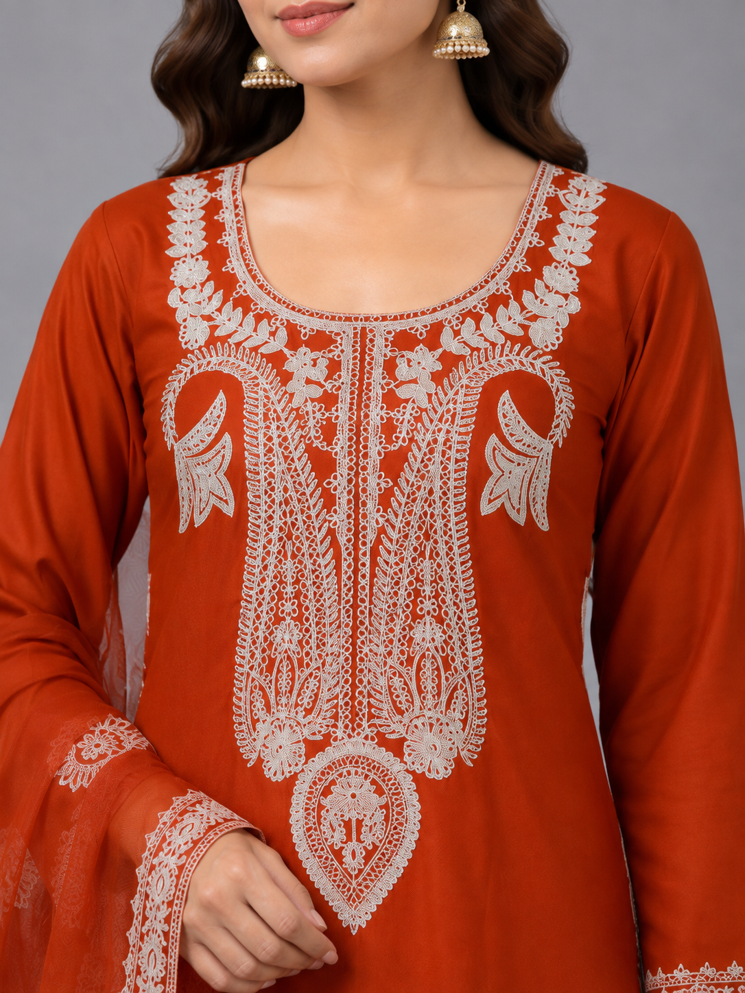 A woman modeling a rust-orange traditional Indian salwar kameez suit with white embroidery and a matching sheer dupatta, set against a solid gray studio background.