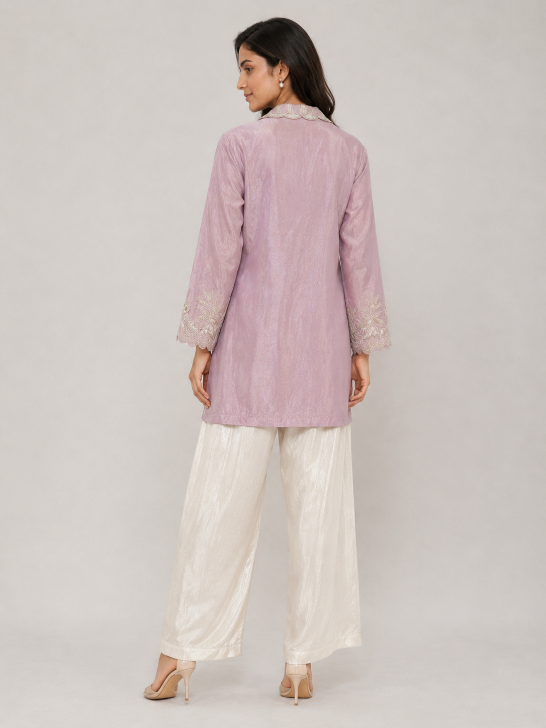 A woman modeling a light pink embroidered kurta paired with white satin palazzo pants and nude heels, set against a solid gray studio background.