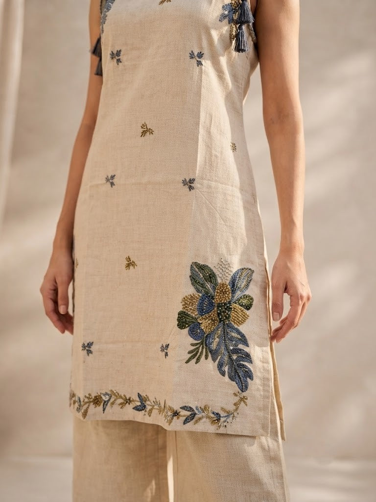 A woman modeling a sleeveless beige kurta with blue and green floral embroidery, paired with matching beige wide-leg pants and navy flats, set against a warm neutral background with soft shadows.