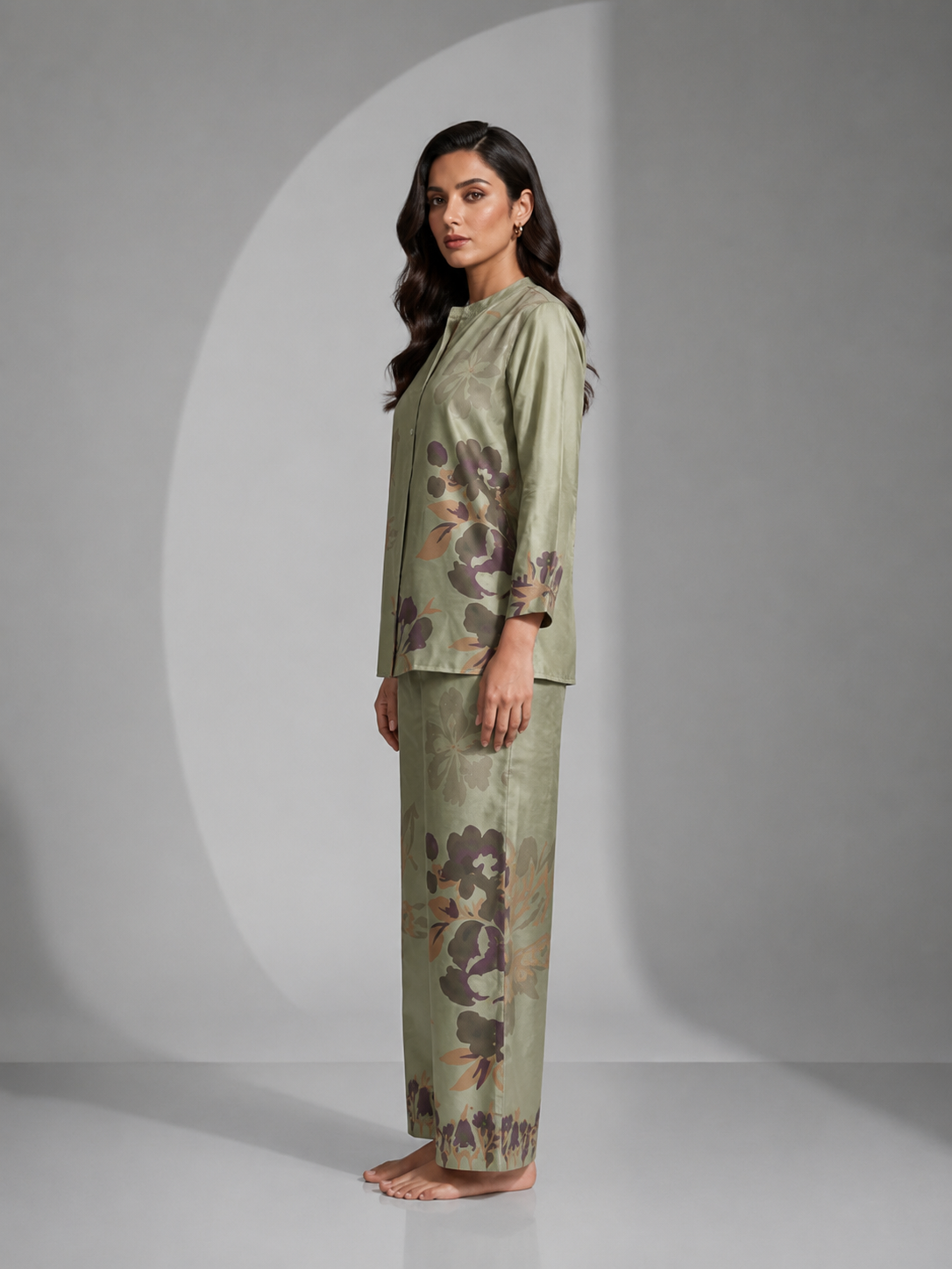 A woman modeling a two-piece satin pajama set in sage green with a floral print, posed against a gray studio background with a circular spotlight effect.